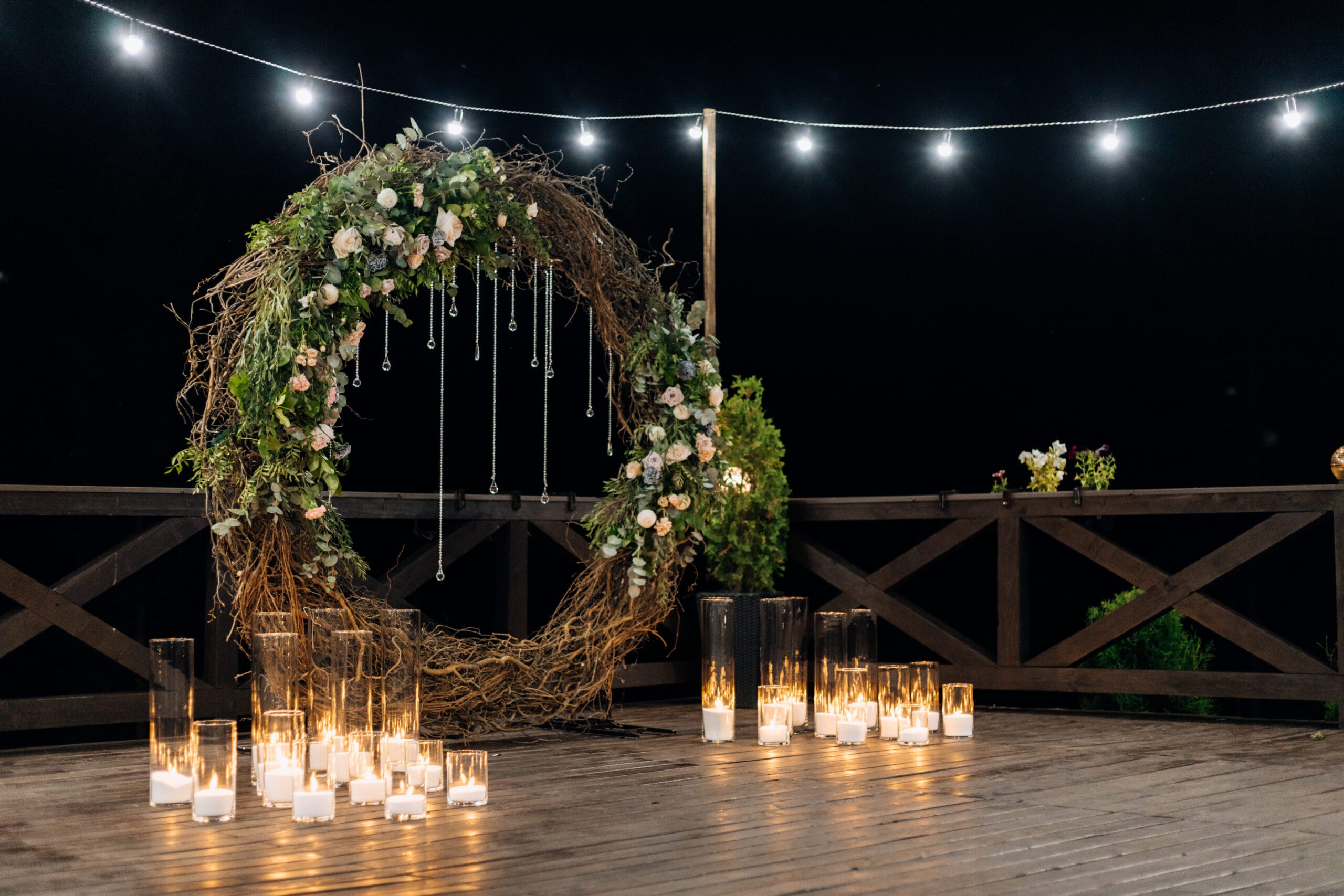 Huge decorative circle made of willow, greenery and pale orange roses with burning candles in the glass candlestick outdoors illuminated in the evening